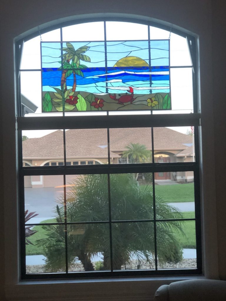 A rectangular leaded stained glass panel depicting a vibrant tropical beach scene. It features two green palm trees on the left, a bright blue ocean with light blue waves, a yellow sun partially submerged in the water, and a red crab on a sandy beach with green foliage and red and yellow flowers in the foreground. The panel is hung within the upper arched section of a large window.