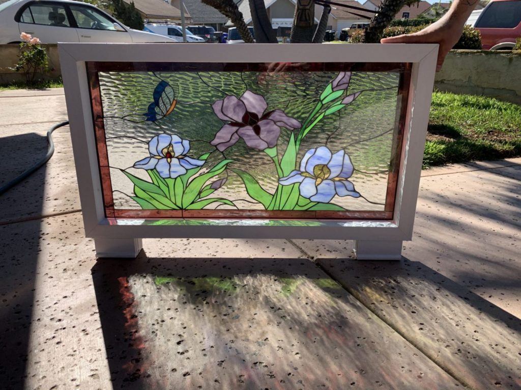 A rectangular stained glass panel in a white wooden frame, depicting a vibrant garden scene. It features three iris flowers (one large purple, two smaller light blue/lavender) with green leaves and stems. A small blue butterfly is visible near the top left. The background is clear, textured glass. The panel stands upright on a sunny concrete surface outdoors, casting colorful reflections and shadows.