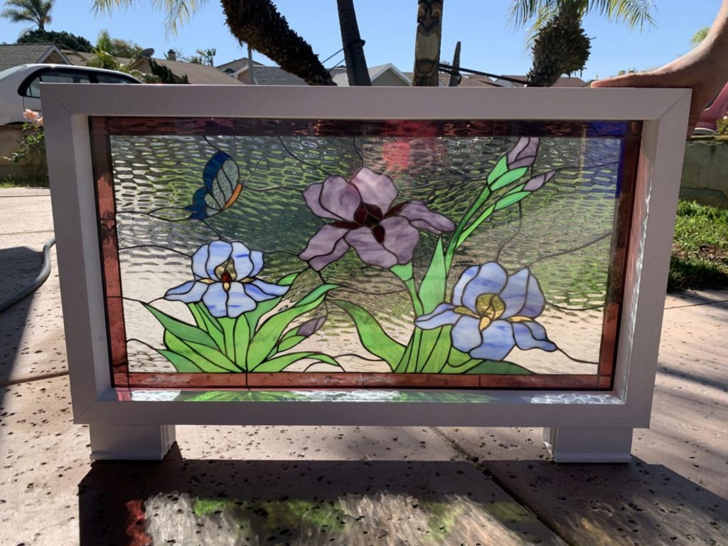 A rectangular stained glass panel in a white frame, depicting a vibrant garden scene with purple and light blue irises, green foliage, and a blue and orange butterfly against a clear, rippled glass background. The panel stands on an outdoor patio surface.