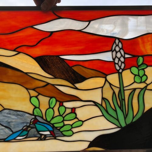 A rectangular stained glass panel showcasing a desert landscape with a bright orange-red sunset sky, rolling tan and brown hills, green paddle cacti, a tall white yucca flower, and two small blue and red birds near a grey rock, all framed in dark metal.