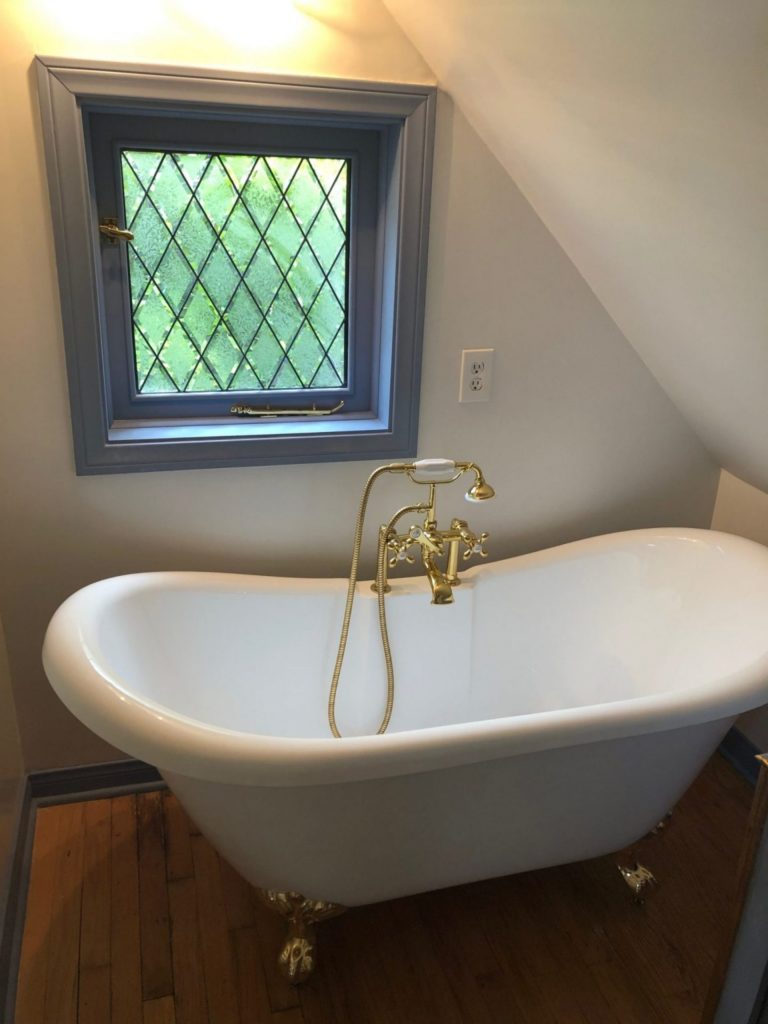 A white clawfoot bathtub with ornate brass fixtures sits below a small window featuring clear, diamond-patterned leaded glass in a blue frame, in a bathroom with sloped attic walls.