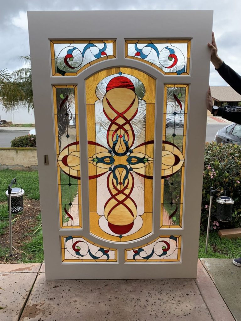 A full-sized, light-colored entry door featuring an integrated stained glass panel. The central design is an ornate, amber-dominant Celtic knot or cross, accented with red and blue. Surrounding smaller panels include clear textured glass and swirling blue, red, and green accents.