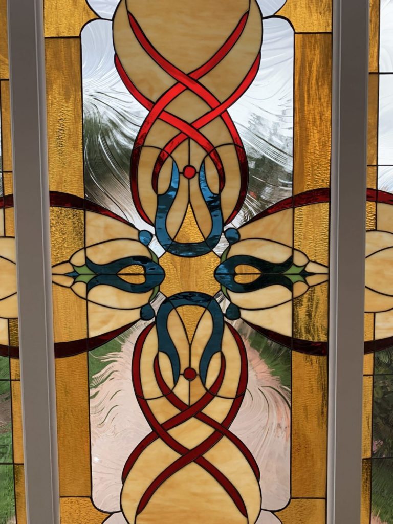 A vertical leaded stained glass door panel featuring a symmetrical, intricate Celtic knot design. The central motif is warm amber glass, flanked by intertwining red ribbons and smaller blue and green decorative accents. Clear textured glass allows light to pass through the background.