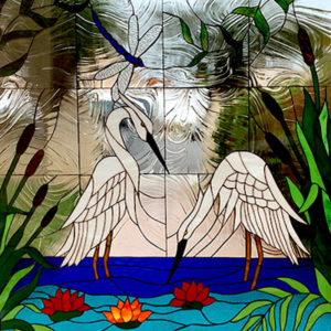 A rectangular stained glass panel depicting a serene wetland scene. Two graceful white egrets face each other, standing in vivid blue water with vibrant red and orange water lilies. A blue-winged dragonfly hovers in the upper left. The scene is framed by lush green cattails and other foliage. The background features clear, highly textured, rippled glass that evokes movement.