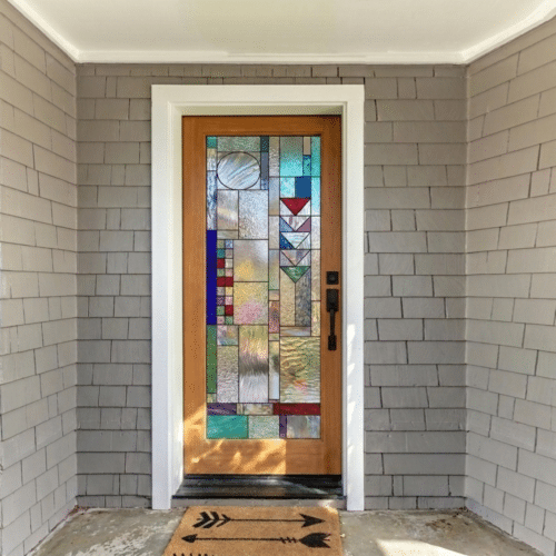 A wooden front door featuring a rectangular stained glass panel with a modern abstract geometric design composed of clear, textured, and colored glass.