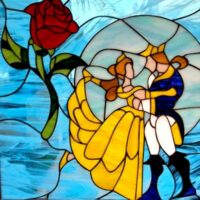 A vibrant leaded stained glass panel depicting a red rose with green leaves on the left, and two figures dancing on the right. One figure wears a flowing yellow gown, and the other a blue and gold coat. The background is composed of clear and wavy light blue glass. The design evokes the Beauty and the Beast fairy tale.
