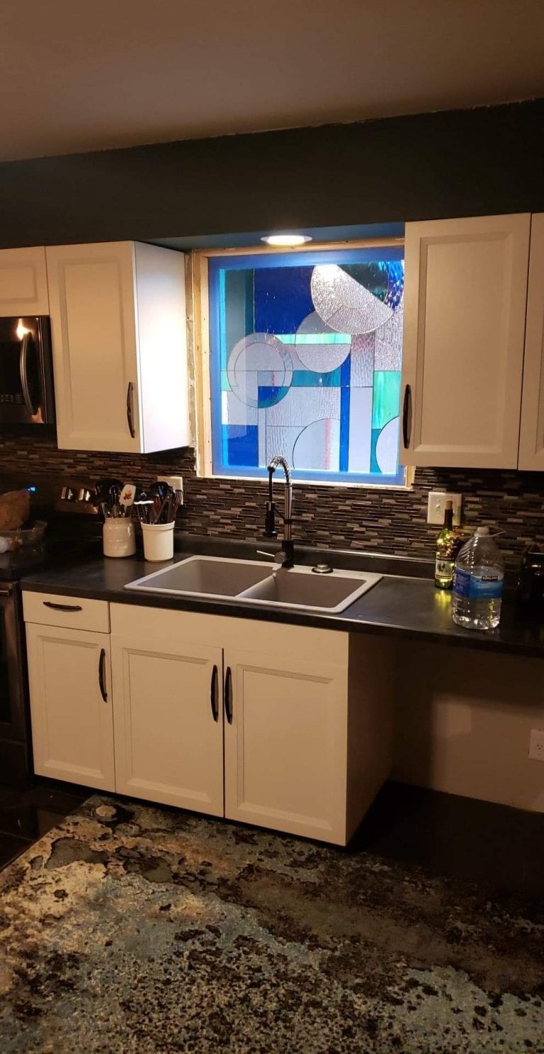 A rectangular, custom-made stained glass window installed above a double kitchen sink, featuring a vibrant abstract geometric design. The panel incorporates various shades of blue, light teal, and clear textured glass, arranged in overlapping circles, rectangles, and linear segments, creating a modern pattern. The window is positioned between white upper kitchen cabinets, above a dark countertop with a mosaic tile backsplash.