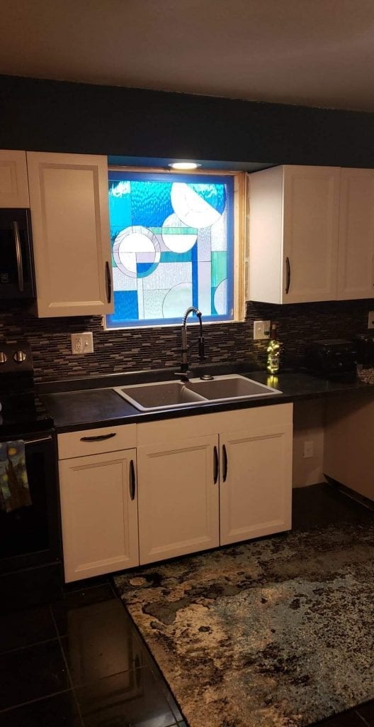 A rectangular stained glass panel featuring an abstract, geometric design with circles, curved lines, and rectangular shapes in various shades of blue, clear, frosted, and lightly textured glass, installed above a kitchen sink with white cabinets.