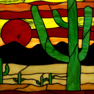 A vibrant stained glass panel depicting a desert landscape at sunset. A large green saguaro cactus dominates the right side, with smaller cacti scattered across the sandy foreground. A prominent red sun is setting behind dark mountains, illuminating the sky with warm stripes of yellow, orange, and red hues.