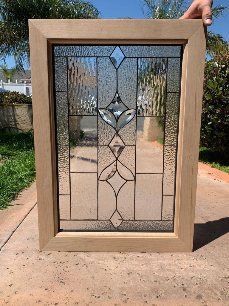 A rectangular clear stained glass panel set in a light-colored wooden frame, showcasing a central geometric motif crafted from multiple clear beveled glass pieces, flanked by various clear textured glass panels.