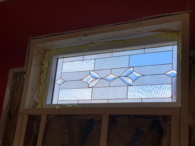 A rectangular, white-framed insulated window featuring a geometric leaded glass panel with clear, various textured, and light blue glass, accented with prominent diamond and leaf-shaped bevels.