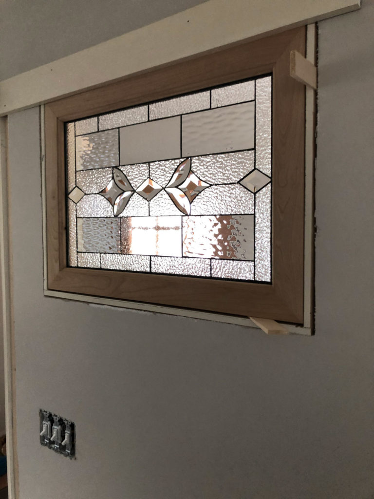 A rectangular clear leaded stained glass panel with a geometric pattern, featuring various textured glass types and central clear bevels, set within a light wood frame during installation in a light grey wall with unfinished trim.