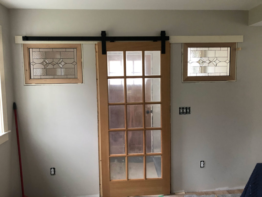 Two rectangular clear stained glass transom panels, featuring a geometric ribbon design with textured and beveled glass, are installed above either side of a light wood French door on a black barn door track. The room has light gray walls, suggesting a renovation or new construction.