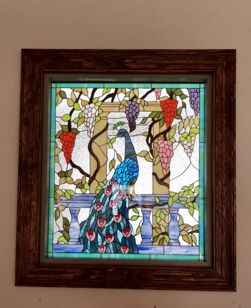 A rectangular stained glass panel in a dark wooden frame, depicting a majestic blue and green peacock perched on a grey balustrade. The peacock is surrounded by grapevines with clusters of red, purple, pink, and white grapes, and light-colored pillars. The background glass is clear and textured.