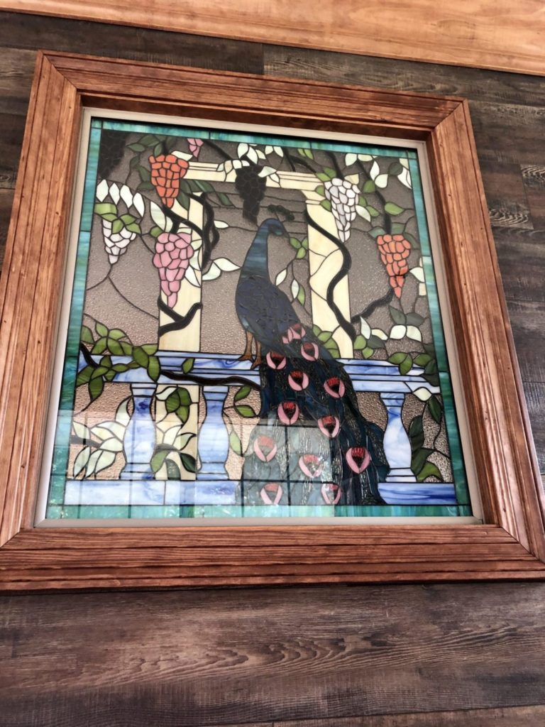 A rectangular stained glass art panel in a rustic wooden frame, depicting a majestic blue peacock with vibrant pink-tipped tail feathers perched on a classical balustrade. Lush green grapevines with clusters of red, pink, and white grapes cascade around a stone pillar, set against a textured, light-filtering background.