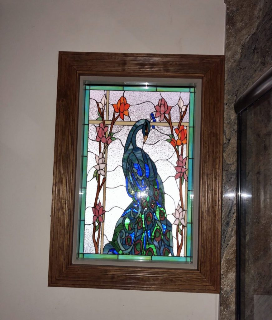 A rectangular leaded stained glass panel featuring a majestic blue and green peacock with intricate feather details, surrounded by pink, orange, and white flowering branches. The background is clear textured glass, all enclosed in a light brown wooden frame.