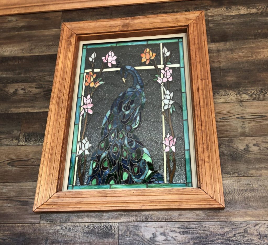 A rectangular stained glass panel in a light wooden frame, featuring a majestic peacock with iridescent blue and green feathers, surrounded by pink, orange, and white floral vines against a textured dark background.