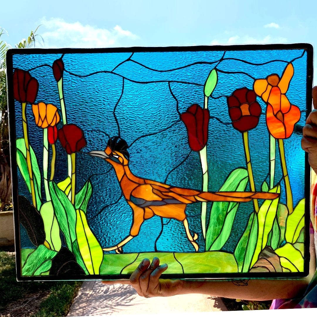 A rectangular leaded stained glass panel held by two hands against a blue sky. The panel depicts an orange, black, and light yellow roadrunner with a crest, striding left to right through green leaves and tall red and orange flower-like plants. The background is a textured bright blue.