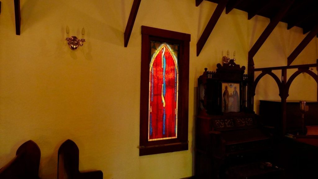 An arched stained glass window with a vibrant abstract design, predominantly deep red with vertical streaks of bright blue and yellow, set in a dark wooden frame within a dimly lit church sanctuary. Wooden ceiling beams, an ornate wooden cabinet with a religious icon, and church pews are visible in the warm interior.