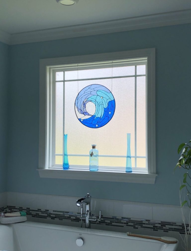 A circular stained glass panel depicting a stylized ocean wave in shades of azure, cerulean, and deep blue, suspended against a frosted bathroom window with white trim. Small clear glass bubbles are integrated into the wave crests, enhancing the fluid design. The panel hangs above a white bathtub in a light blue room.