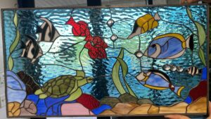 A rectangular stained glass panel featuring a vibrant underwater scene. Three tropical fish in blue, yellow, and black-and-white, a green sea turtle, and red coral are set against a background of textured blue water. Green seaweed and clear glass bubbles add detail, while the bottom depicts a rocky seabed in brown, pink, and blue hues. The panel has a dark metal frame and is being held by hands.