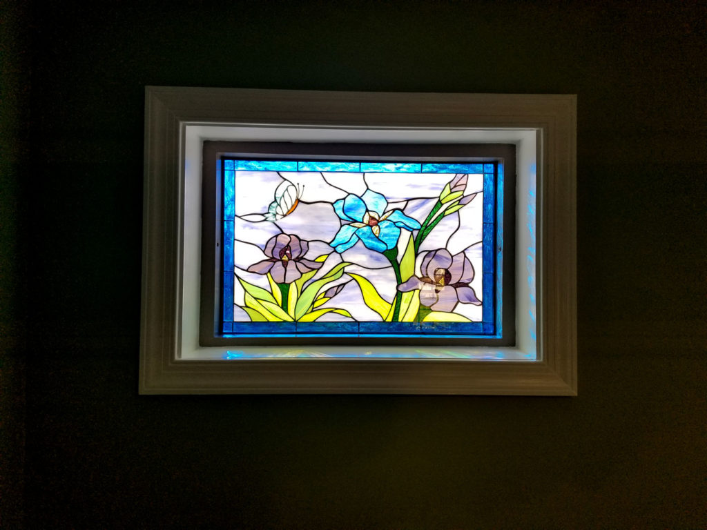 A rectangular leaded stained glass panel depicting vibrant blue and soft purple irises with green leaves and a small butterfly, set within a white frame against a dark wall.
