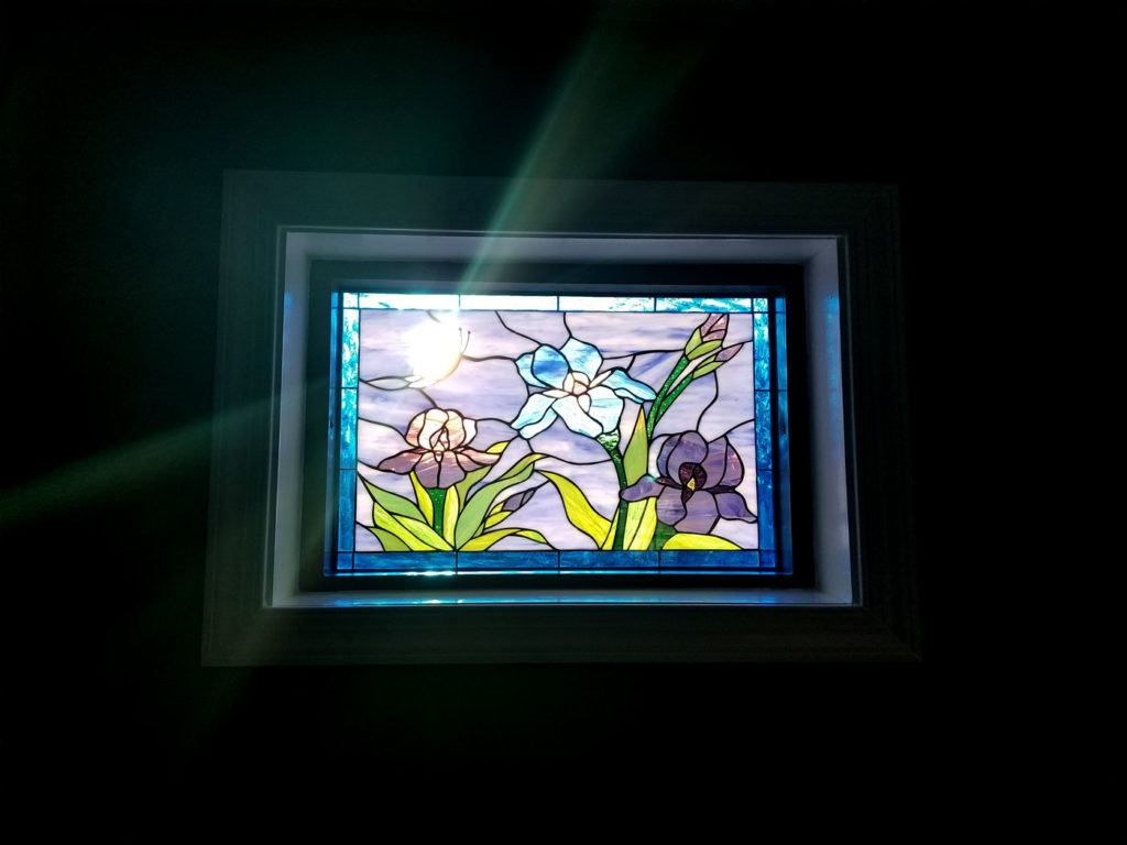 A rectangular stained glass window panel depicting three vibrant iris flowers: one white, one lavender, and one deep purple, with green leaves and stems against a light blue background. A bright, sun-like circle is visible in the upper left, with rays of light passing through the glass and window frame, casting a luminous effect in a dark room.