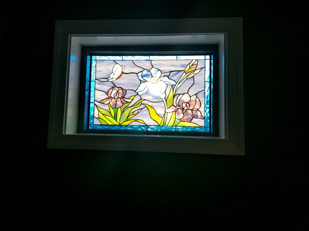 A rectangular stained glass window panel depicting a vibrant floral scene. It features a central white iris, flanked by two pinkish-purple irises, with green and yellow foliage. A white butterfly is visible in the upper left corner. The background is a mix of light blue and lavender swirled glass, all bordered by a rich blue glass frame. The panel is set within a white window frame against a dark wall.