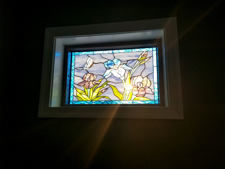 A rectangular leaded stained glass panel featuring a vibrant floral scene. Three irises with white, pink, and lavender petals, along with green leaves and stems, are depicted against a light blue and purple sky background. A delicate white butterfly flies in the upper left, and the bottom border has a light blue water ripple design. The panel is set in a white window frame, with a prominent ray of sunlight shining through and illuminating the central iris.