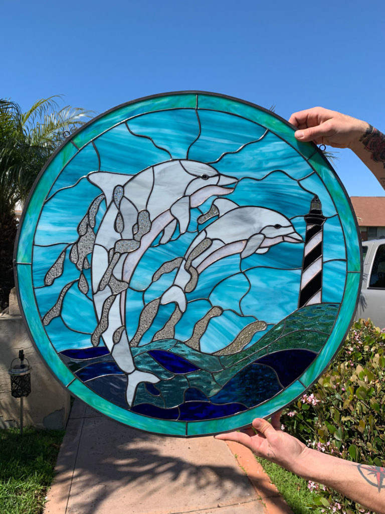 A round stained glass panel featuring two white and gray dolphins leaping from blue textured water, with a black and white striped lighthouse on a dark green island under a clear blue sky. The panel is leaded with a dark frame.