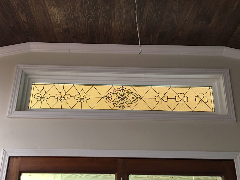 A rectangular clear and textured stained glass transom window installed above a wooden door. The leaded design features three fleur-de-lis symbols on the left, a central ornate floral medallion, and three shamrocks on the right, all against a clear diamond-patterned background. The transom is framed with white molding, with a dark wood plank ceiling visible above.