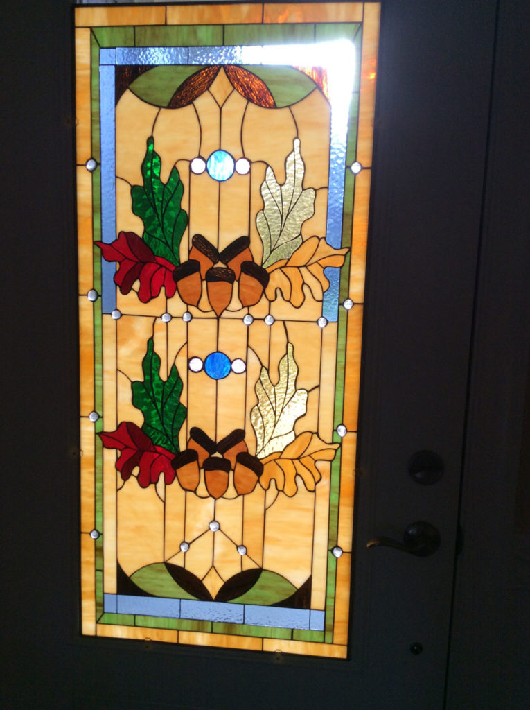 A rectangular stained glass panel featuring a symmetrical autumnal design with clusters of green, red, and golden oak leaves and brown acorns, set against a warm, textured amber background. The panel is adorned with clear beveled jewels and blue circular accents, bordered by geometric patterns in green and blue.