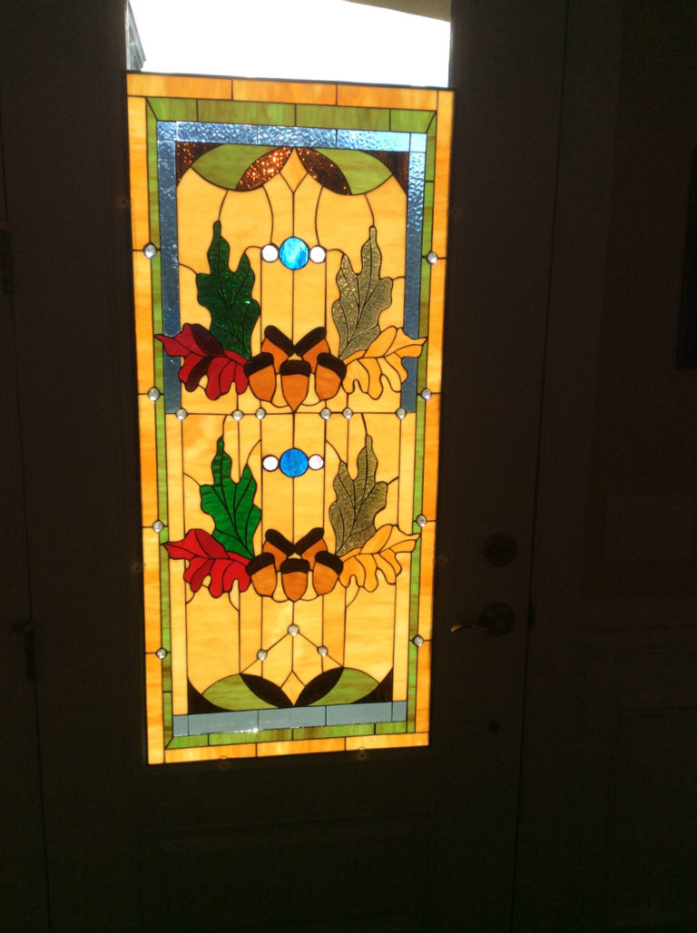 A rectangular stained glass door panel featuring a symmetrical fall-themed design with vibrant red, green, and golden oak leaves, clustered around brown acorns. The background is warm amber-yellow, accented with two bright blue circular jewels, textured clear glass, and a green border. Sunlight illuminates the panel installed in a white door.