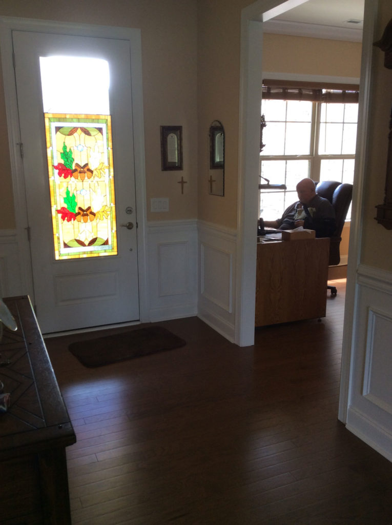 A rectangular stained glass panel in the upper half of a white front door, depicting an autumn design with leaded acorns and colorful red and green oak leaves against a textured amber glass background. The panel allows natural light into a home entryway.