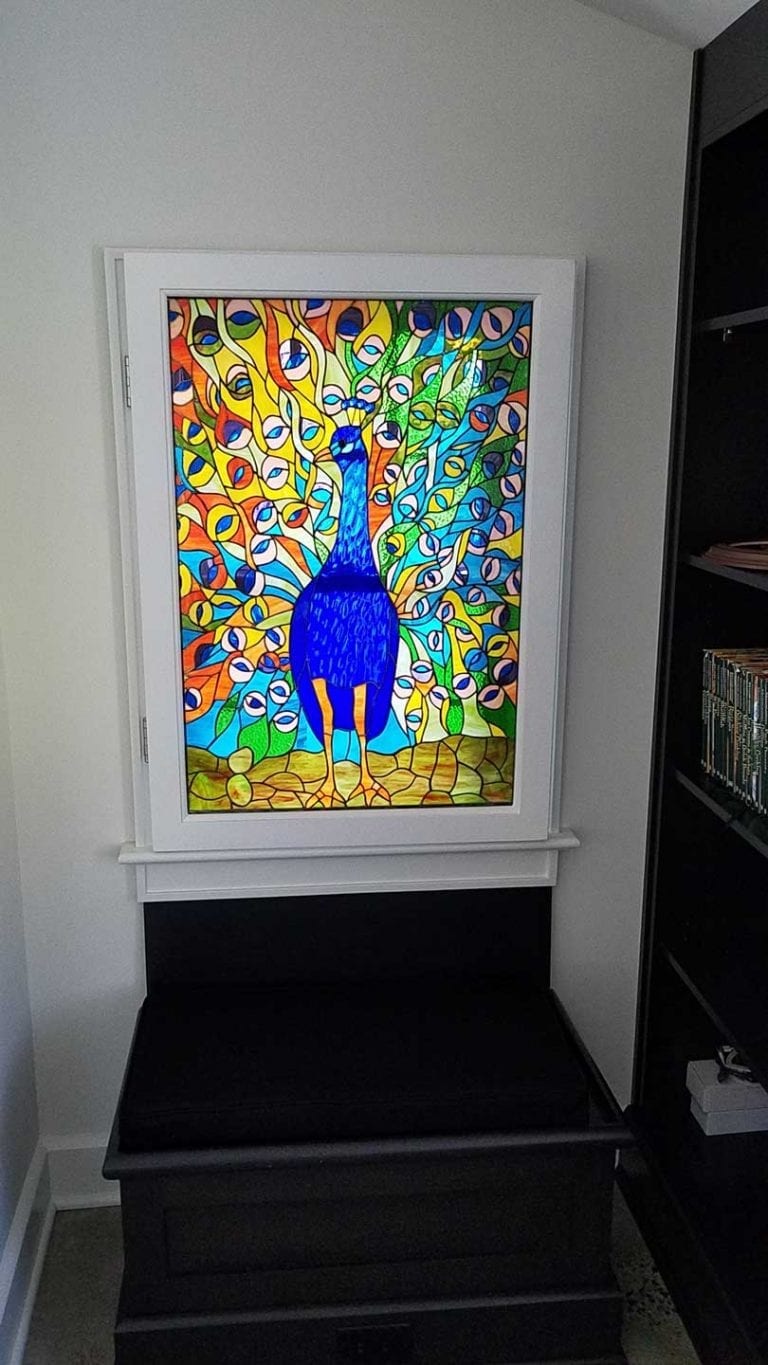 A rectangular stained glass panel featuring a vibrant blue peacock with its tail fanned out in a colorful mosaic of green, yellow, orange, and blue glass. The panel is set in a white frame above a dark storage bench.