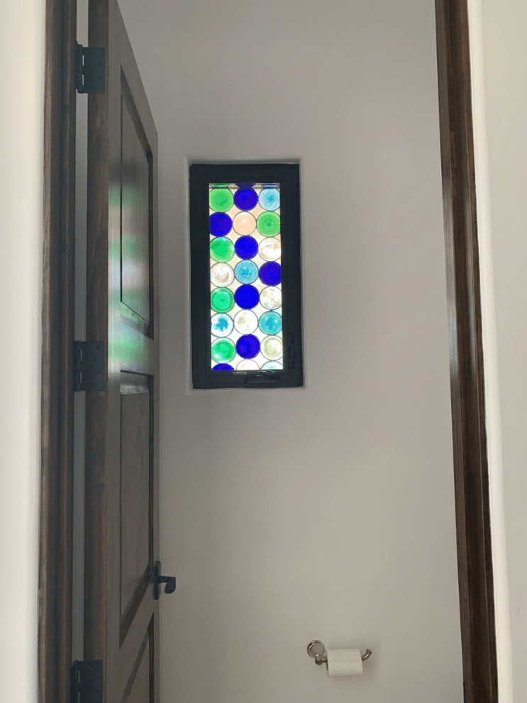 A vertically-oriented rectangular stained glass window in a dark frame, featuring a dense pattern of blue, green, and clear glass rondels.