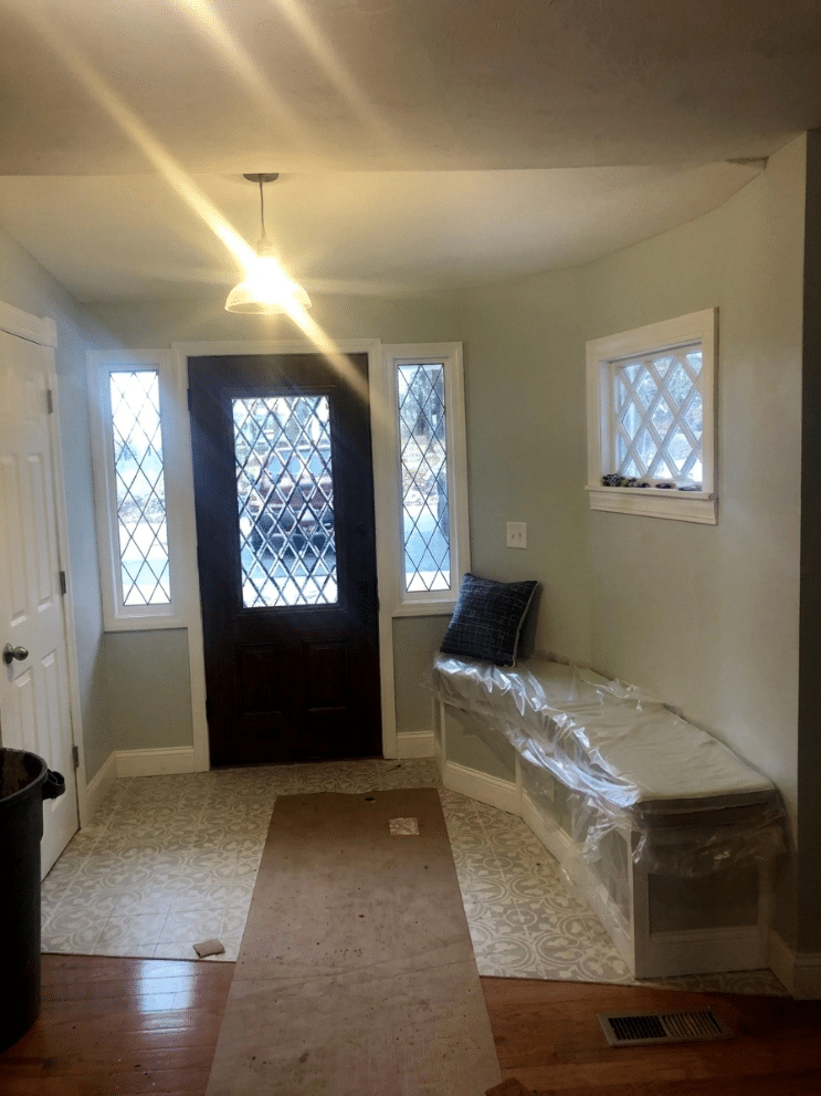 A dark brown entrance door with clear diamond pattern leaded glass, flanked by two matching narrow sidelight windows. On the adjacent wall, a rectangular window with the same clear diamond pattern is visible, above a white built-in bench.