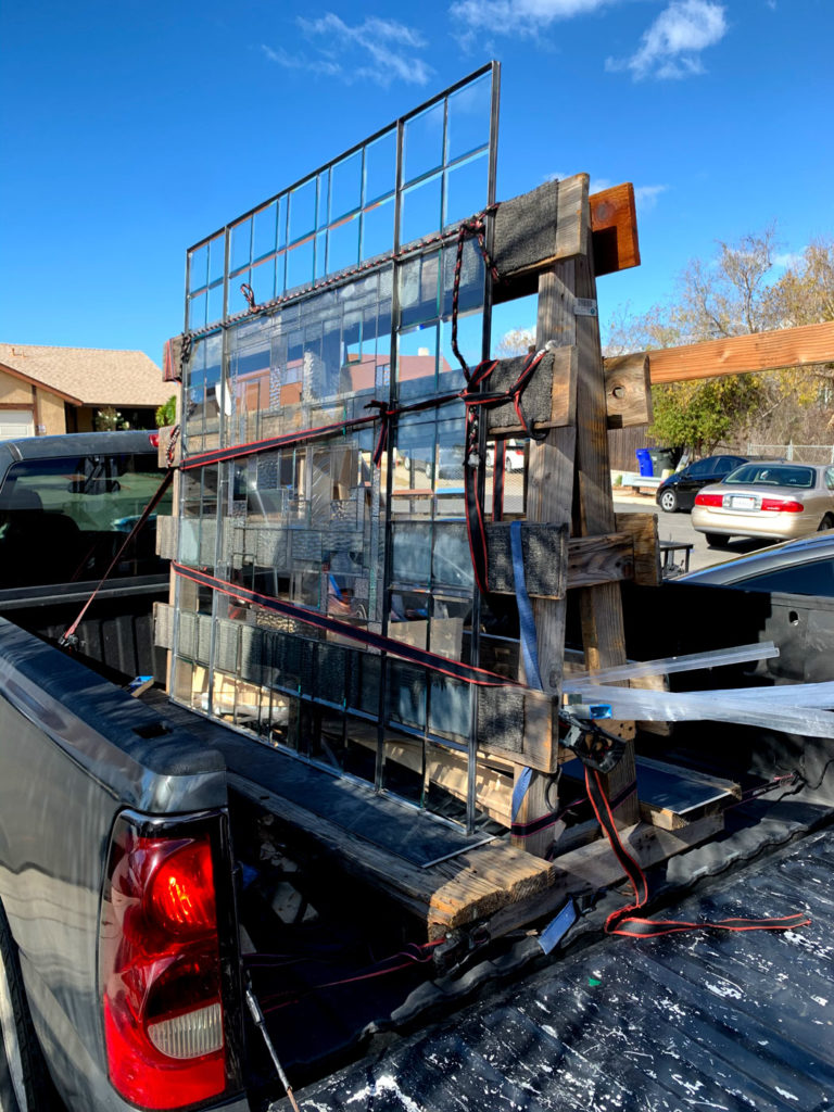 Multiple large, clear leaded stained glass window panels, securely strapped onto a custom wooden frame in the back of a dark gray pickup truck. The panels feature a simple grid pattern, reflecting the clear blue sky and residential buildings in the background.