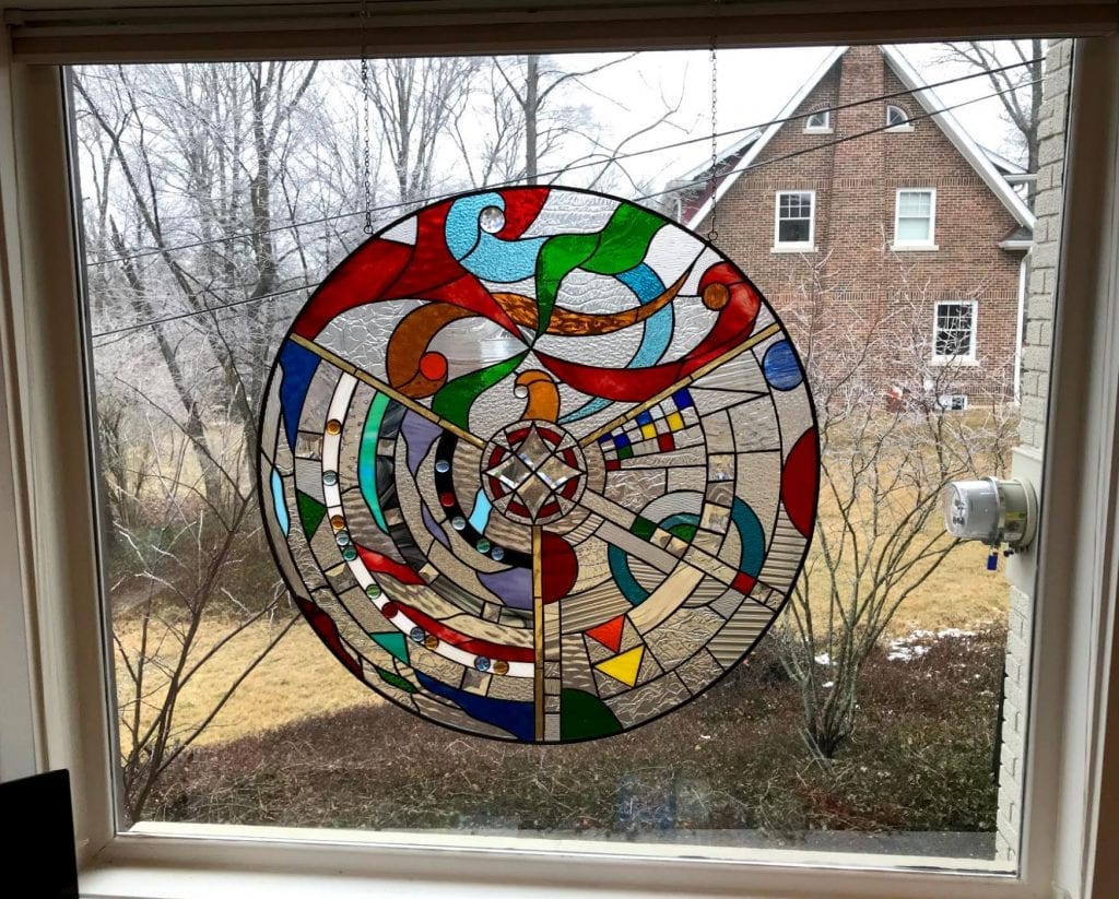 A large, round leaded stained glass panel with a vibrant abstract geometric design. It features a central clear beveled starburst surrounded by swirling patterns in rich reds, blues, greens, amber, yellow, and clear textured glass.