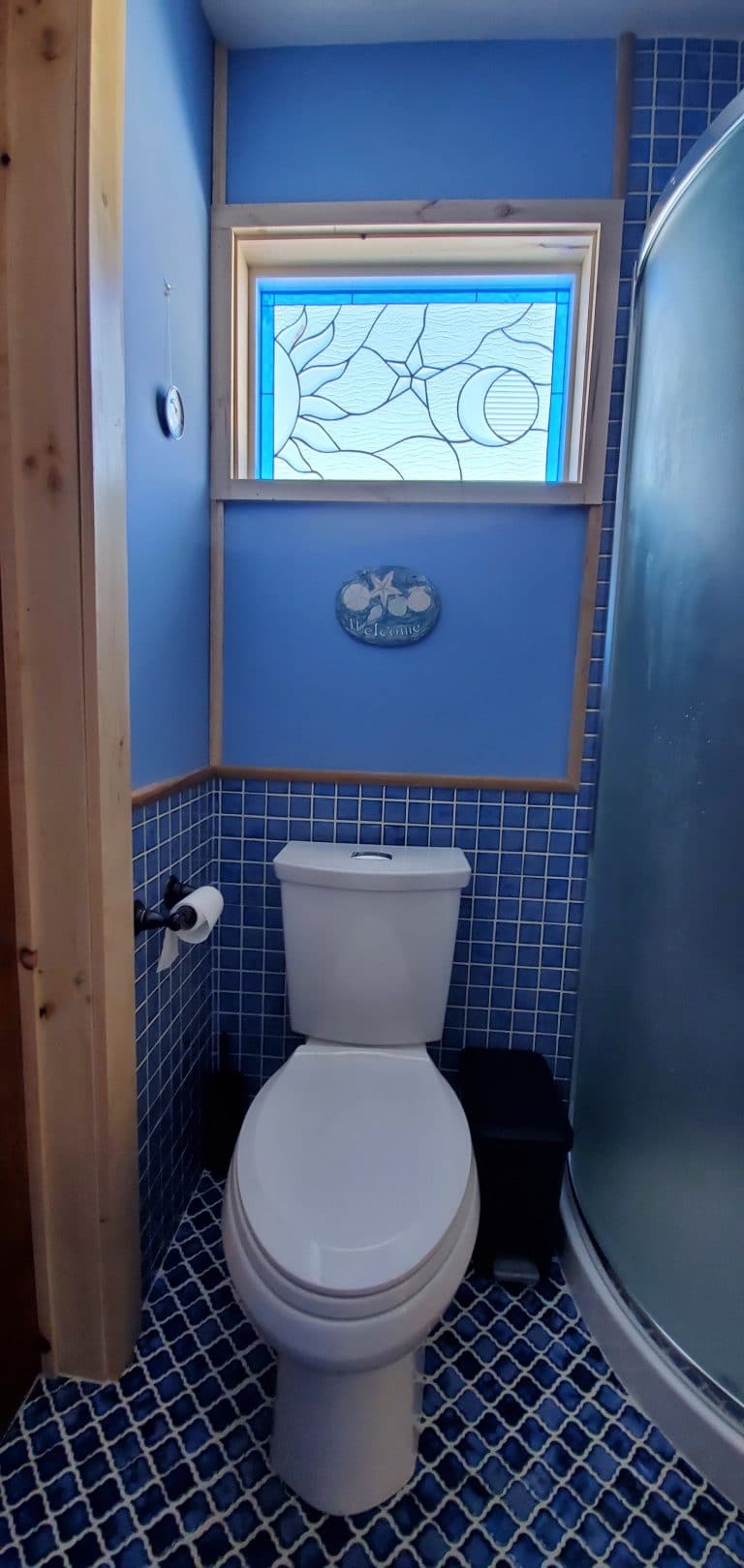 A rectangular stained glass window featuring a celestial design of a stylized sun, star, and crescent moon, set within a light blue border and predominantly clear textured glass, installed above a white toilet in a blue-tiled bathroom.