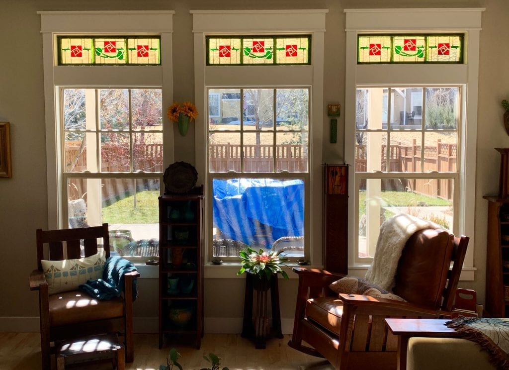 Three identical rectangular stained glass transom panels, each featuring a repeating stylized red rose with green leaves on a light amber background, leaded and installed above clear double-hung windows in a bright living room.