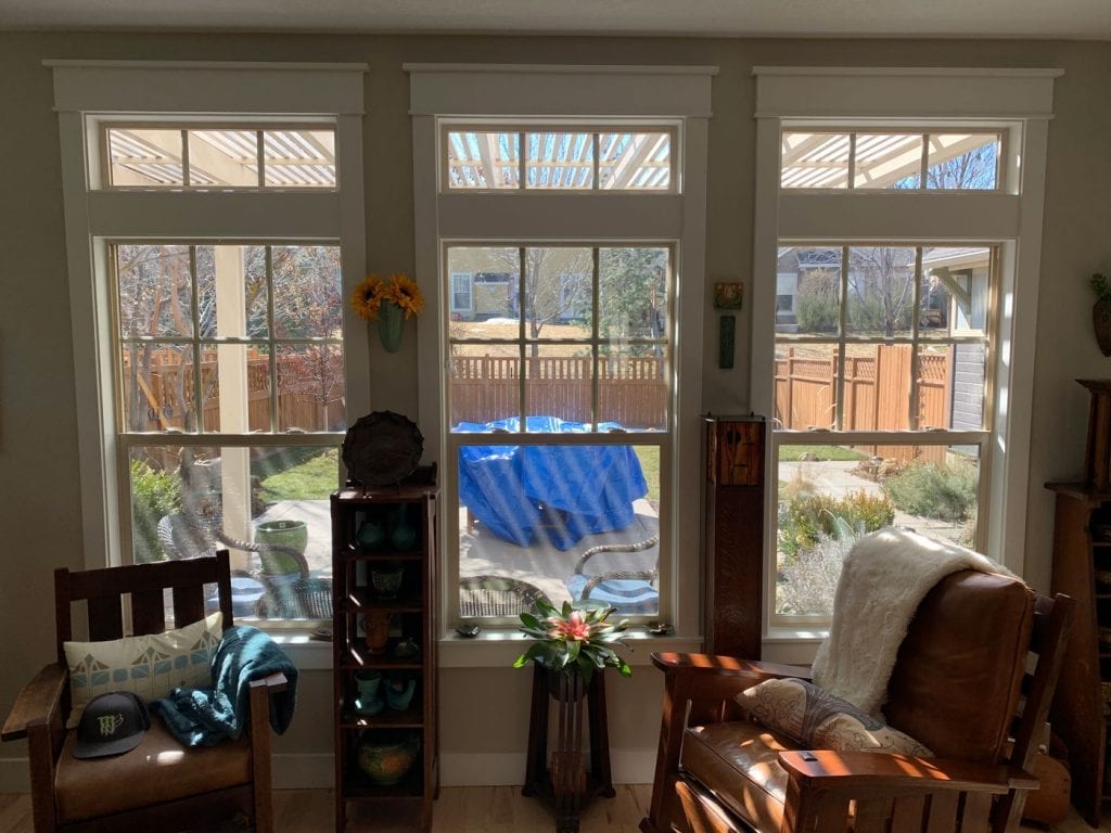 A bright living room featuring three tall, white-framed residential windows, each with an upper transom, looking out onto a sunny backyard. The windows offer ample natural light and a view of a wooden fence and greenery, ideal for custom stained glass.