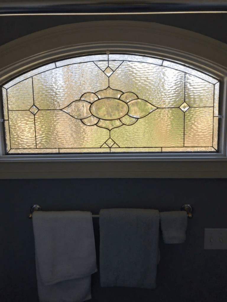 An arched, clear leaded stained glass panel featuring a central stylized floral medallion and geometric diamond accents, set in a window above a bathroom towel rack.