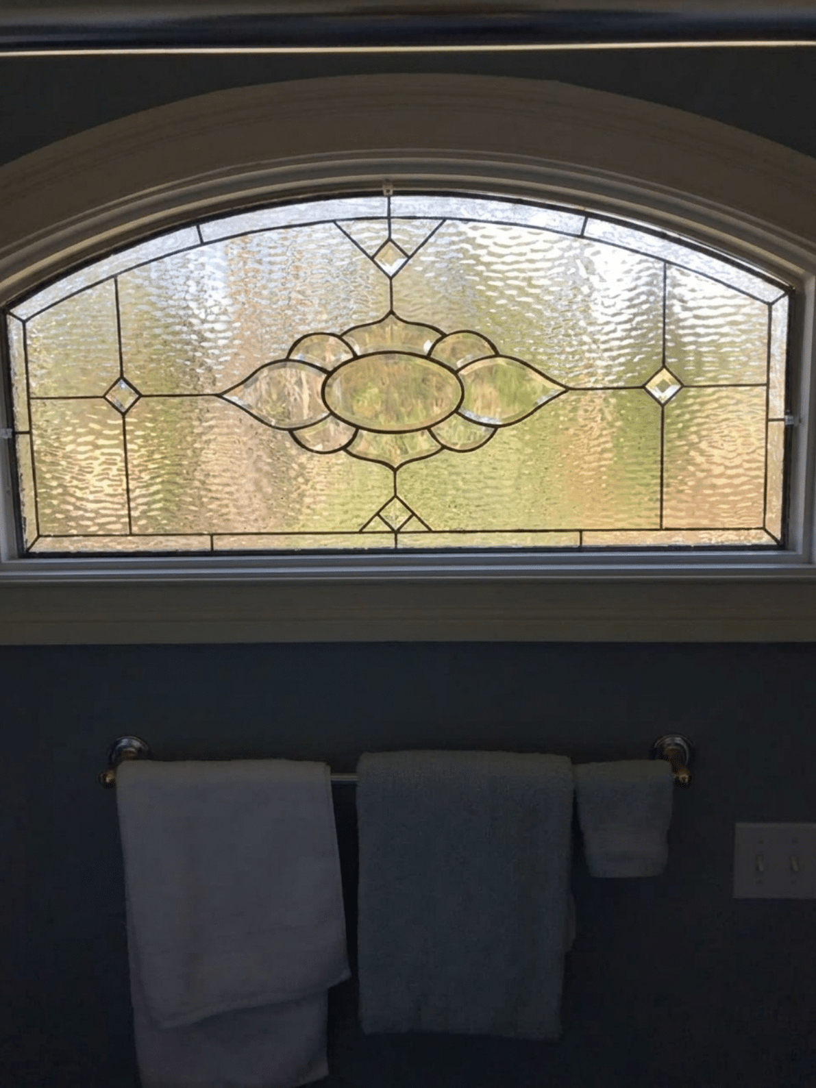 An arched, clear leaded stained glass panel featuring a central stylized floral medallion and geometric diamond accents, set in a window above a bathroom towel rack.