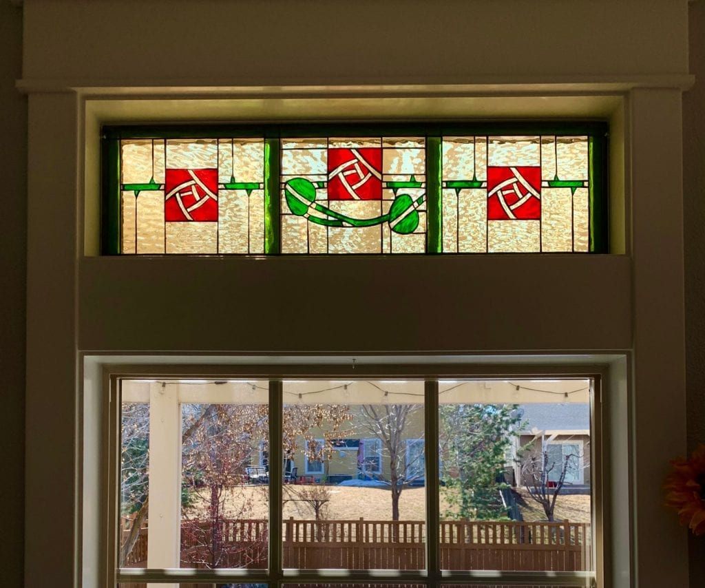 A rectangular stained glass transom window featuring a striking Art Nouveau-inspired design. Three vibrant ruby red squares, each with a white geometric stylized rose or knot pattern, are centered within textured clear glass panels. These squares are interconnected by horizontal emerald green bars and a graceful, flowing green vine with two leaves. The entire panel is framed in a dark lead came, set against a light-colored wall with a larger window below showing a winter backyard scene.