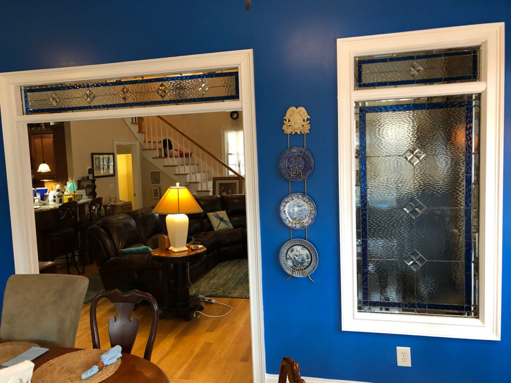 A home interior featuring two custom leaded stained glass installations against vibrant royal blue walls. On the left, a long, rectangular transom panel is installed above an interior doorway. It showcases clear textured glass, multiple diamond-shaped clear bevels, and a striking cobalt blue border. To the right, a matching vertical rectangular window panel is set into the wall, repeating the clear textured glass, clear bevels, and cobalt blue border design. Both panels are framed by clean white trim. The doorway offers a view into a brightly lit living room with a brown leather couch and an adjacent kitchen area.