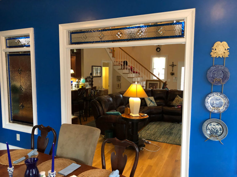 Two interior stained glass panels, framed in white, against a bright blue wall. The taller rectangular panel and the wider transom panel over an entryway both display a leaded geometric pattern using clear, heavily textured glass for privacy, accentuated with deep blue glass borders and diamond shapes.
