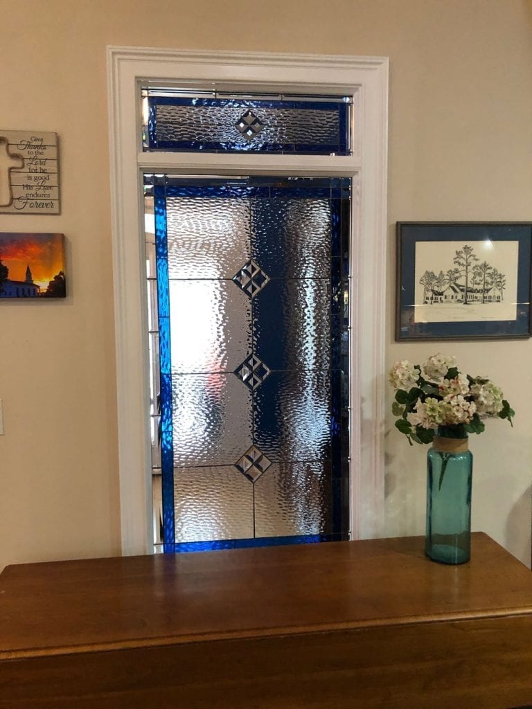 A rectangular leaded stained glass panel with a vibrant sapphire blue border and clear, heavily textured glass in the main sections. Small clear beveled diamond shapes are strategically placed within the geometric pattern, and the panel includes a distinct upper transom-like section, all set in a white window frame.
