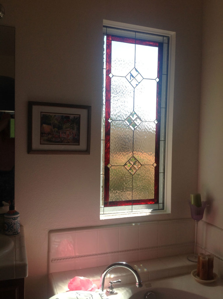 A vertical rectangular leaded stained glass window, featuring clear textured glass for privacy, a rich red border, and three central clear beveled diamond medallions. The window is set in a light-colored bathroom above a bathtub.