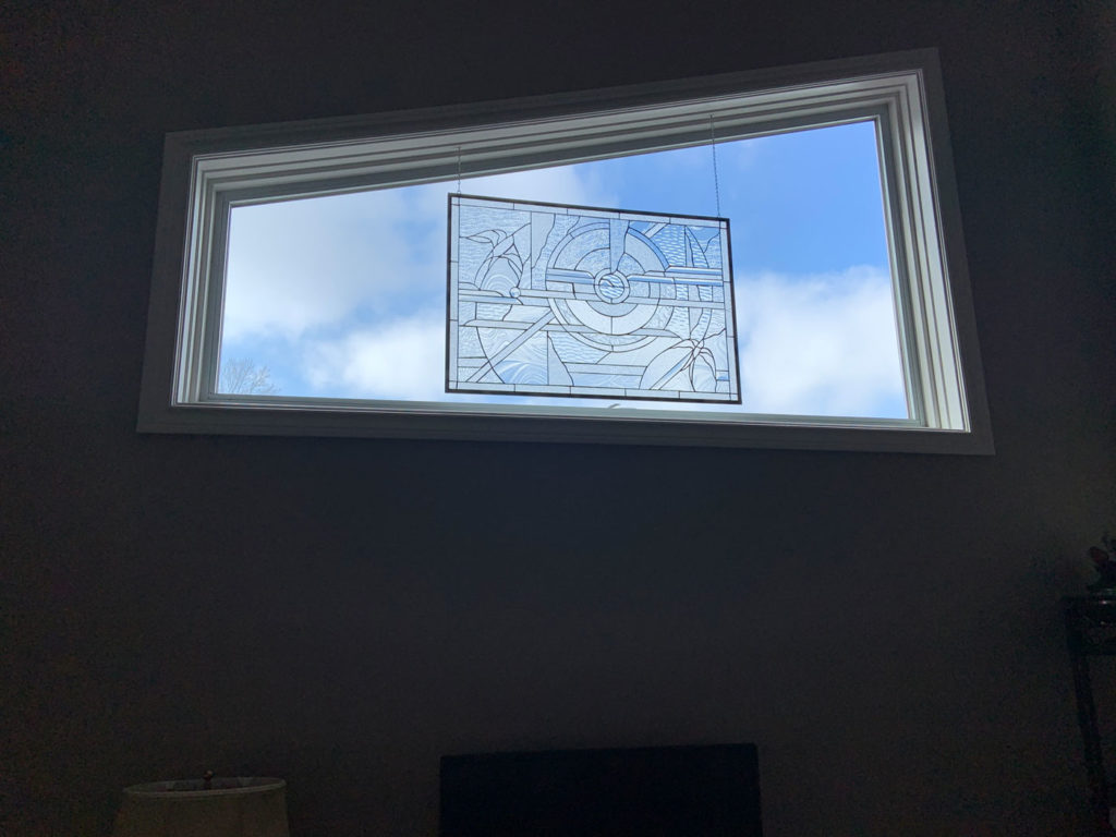 A rectangular clear textured stained glass panel with lead lines forming an abstract, geometric pattern, hanging from chains in a white-framed window against a bright blue sky with white clouds.